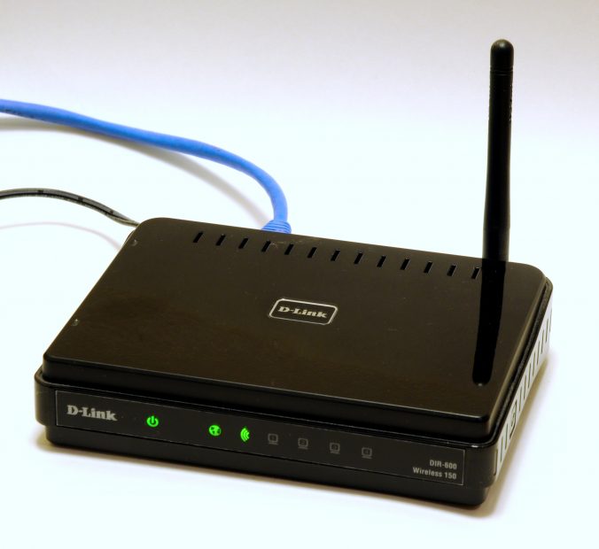 En bild som föreställer en svart router av märket D-Link. Routern har en svart antenn och den är ansluten med en blå nätverkskabel samt strömkabel. Tre gröna indikatorlampor är tända.